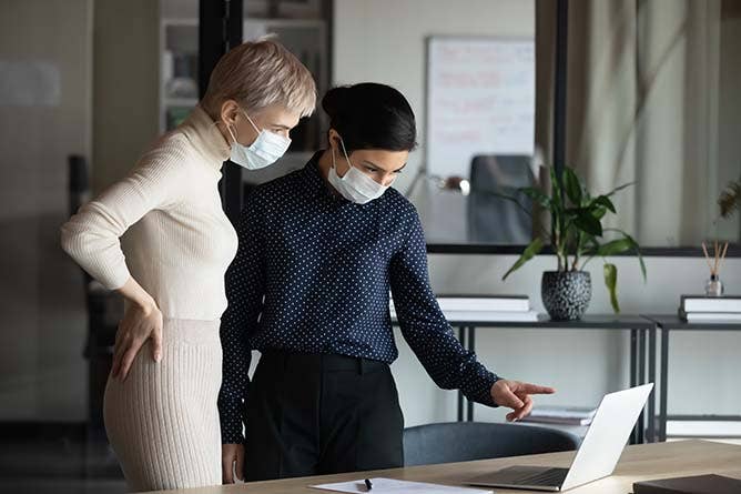 Focused two young business women in medical facial protective masks standing near table, looking at laptop screen, discussing project details