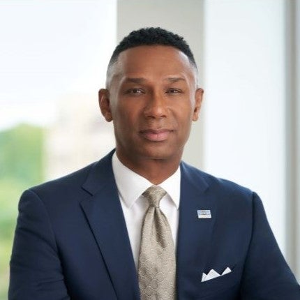 Head shot of Johnny C. Taylor, a black man with black hair wearing a dark suit and gray tie with windows as a back drop.