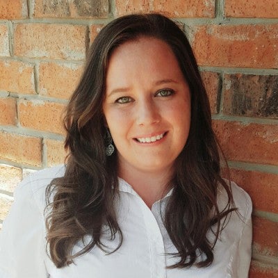 Rachael is a white woman with long brown hair. She is smiling and wearing a white shirt with silver earrings and standing in front of a brick wall backdrop.