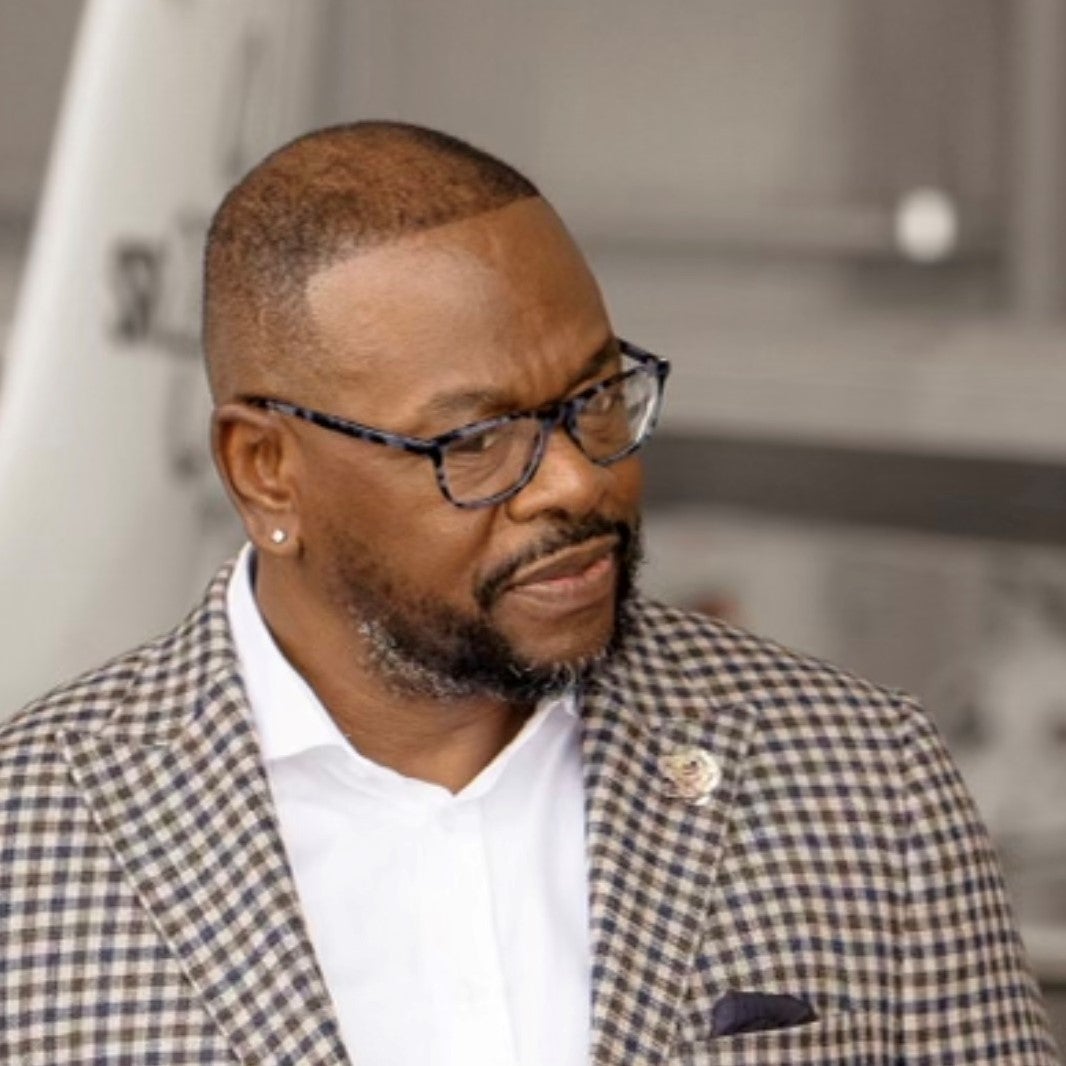 Milton Collins is a black man wearing a checkered suit and black glasses.