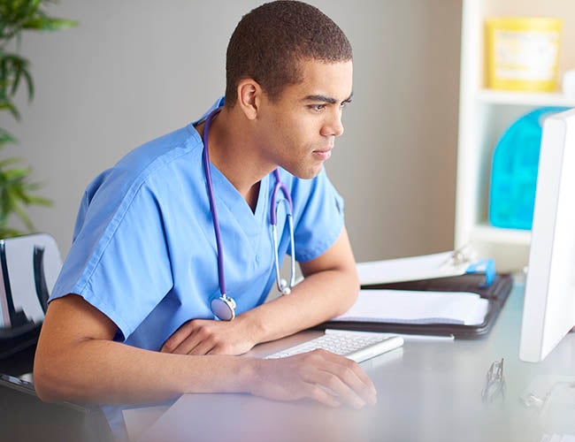 male nurse or doctor sits at his desk looking at his computer screen and writing in his pad . Sunlight pours across the desk from the window beside the desk. He is wearing scrubs .