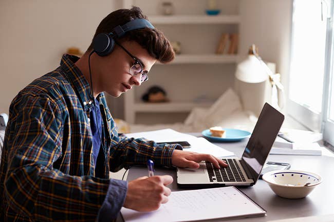 Teenage student wearing headphones and sitting at laptop with notebook