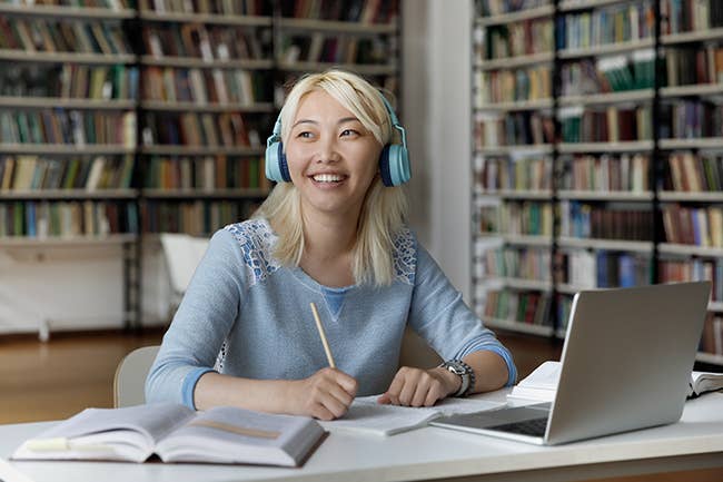 Teenage student wearing headphones in library working at laptop