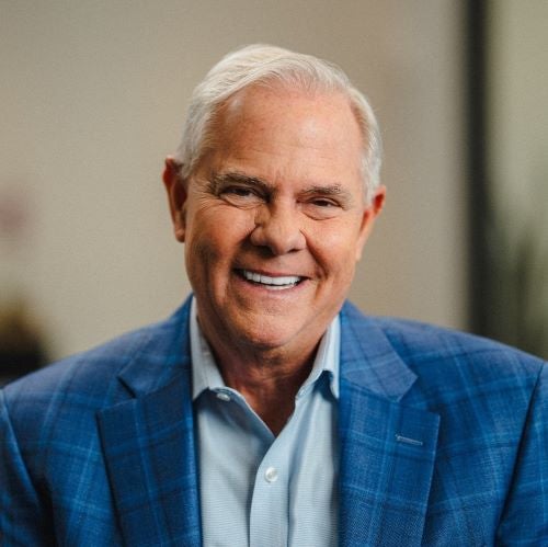 David Simmons is a white man with gray hair and is smiling, wearing a light blue button up shirt and a dark blue plain suit coat.
