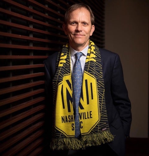 John Ingram is a white man with brown hair, wearing a Nashville Soccer Club scarf over a suit and tie, leaning against a brick wall and smiling.