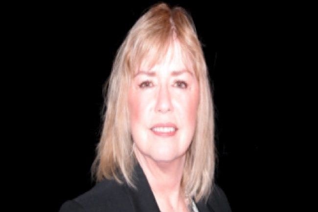Marti Garlett is a white woman with blond hair and is pictured smiling and wearing a dark business suit against a black background.