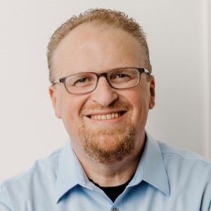Casey Clark is a white man with red-blond hair and glasses. He is smiling and wearing a blue button down shirt.