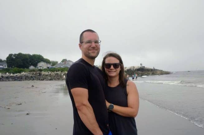 Jake and Sammi Piispanen stand smiling on a beach on an overcast day. They are a white couple in swimwear and both wearing glasses.