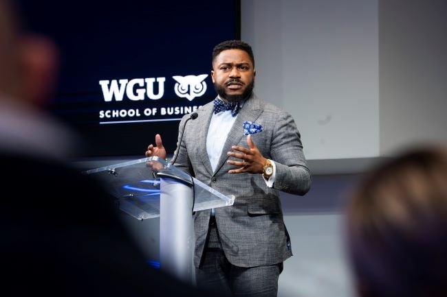 Brandon Fleming is a black man wearing a grey suit and standing at a speaker's podium addressing a crowd.