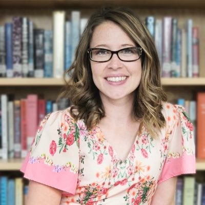 Carley Maloney is a white woman with brown hair and glasses. She's wearing a pink floral blouse and sitting in front of a bookcase.