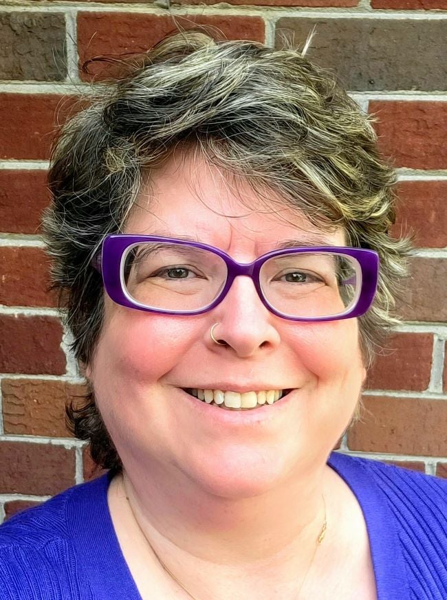 Nicole Buchanan is a white woman with short salt and pepper wavy hair. She is wearing a royal blue shirt with purple glasses and is standing against a brick background.