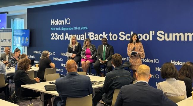 A panel of 4 presters sit on a stage in front of an audience at Holon IQ