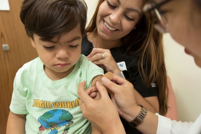 A young boy in a green shirt sits in his mother's lap while a medical provider puts a bandage over a recent immunization