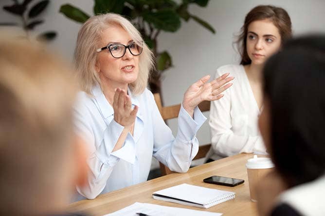 Attractive aged businesswoman, teacher or mentor coach speaking to young people, senior woman in glasses teaching audience at training seminar, female business leader speaker talking at meeting; Shutterstock ID 1043108527; PO: 123