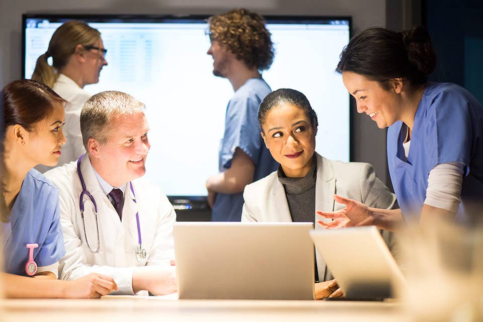 Asian woman nurse, older white man healthcare professional, African American woman healthcare professional, and white woman nurse looking at an laptop computer.  Two healthcare professionals are talking in the background.  This logo is used on the Management Jobs in the Healthcare Industry page.