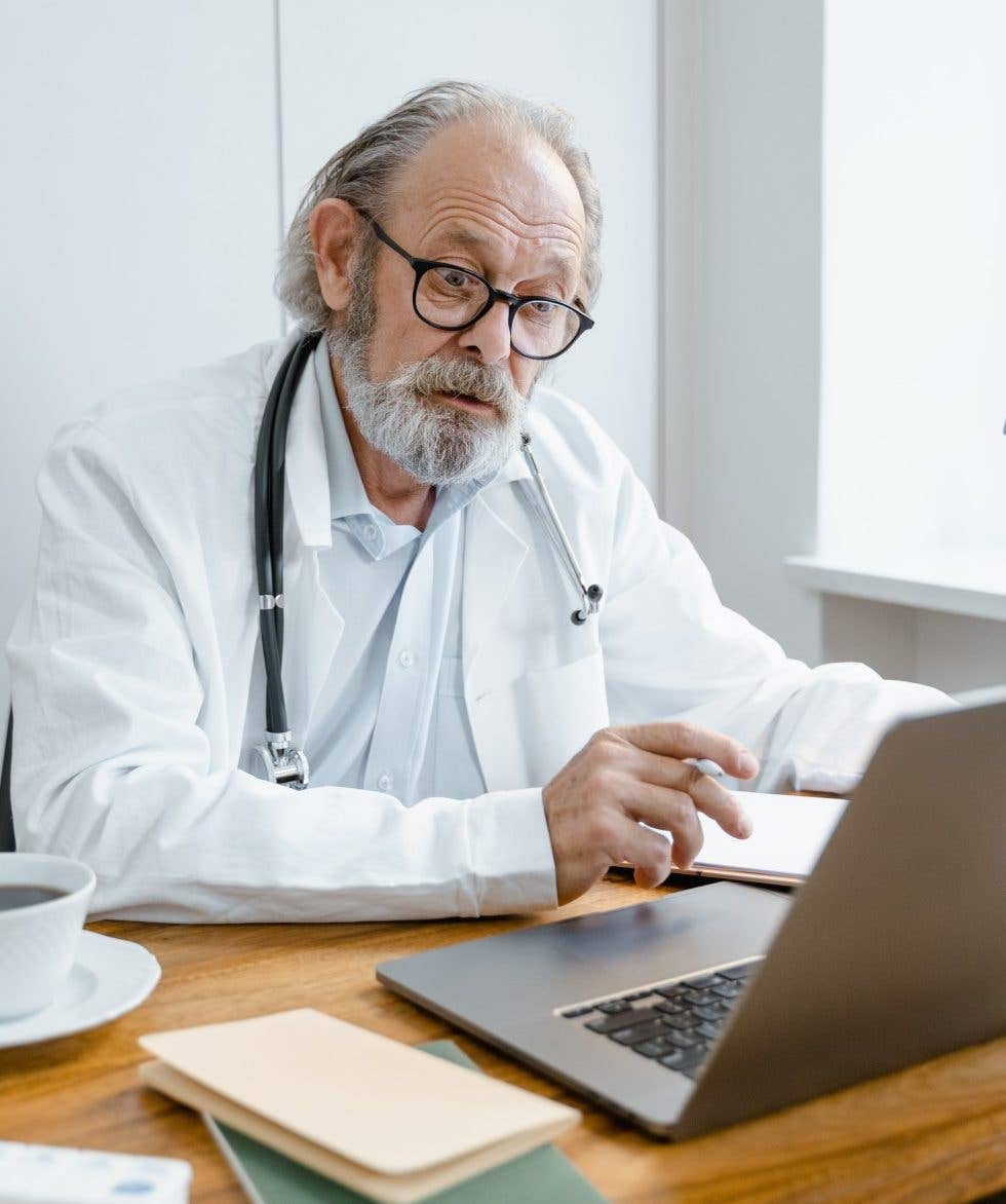 Male doctor with gray hair and beard talking to someone on laptop video call