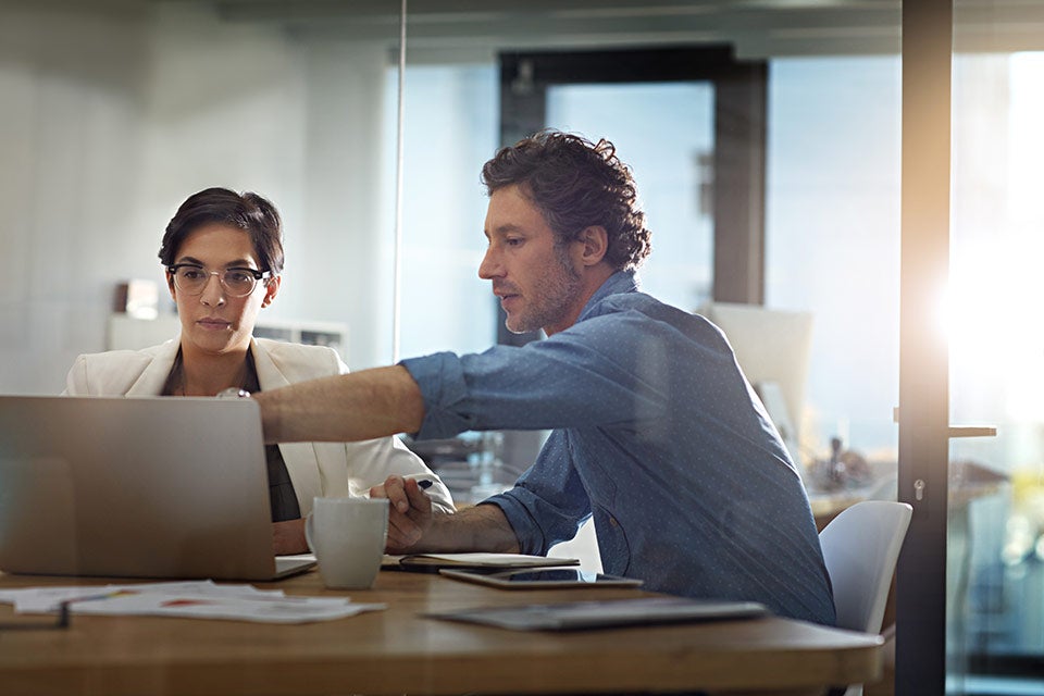 Hispanic business woman and white business man working on an Apple laptop computer.  There are papers, a tablet computer, a coffee cup on the table.  These professionals appear to be in a business environment.
