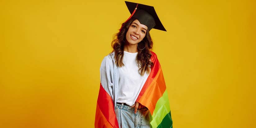 A smiling student wearing a graduation cap and draped in a rainbow flag. 