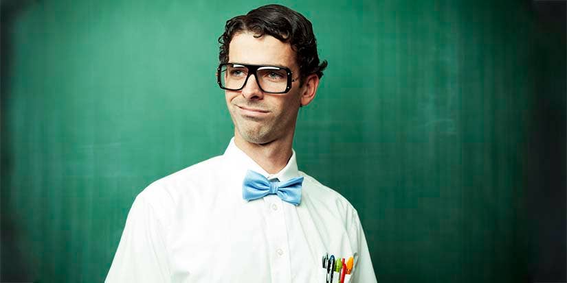 A man dressed with a bow-tie, thick framed glasses, and sporting a pocket protector stands in front of a blackboard.