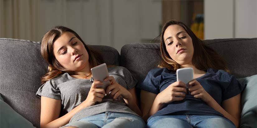 Two young girls slouch on a couch, staring at their phones.