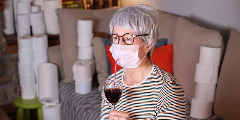An older woman sips wine in her house surrounded by toilet paper rolls with a mask on.