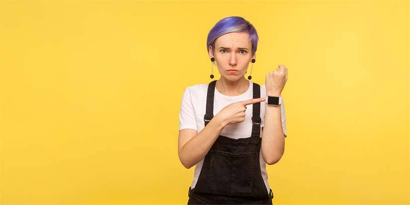 A young girl against a yellow background points impatiently at her smart watch.