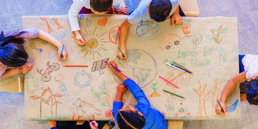 A group of children  surround a table with the world and various crayon drawings all around it.