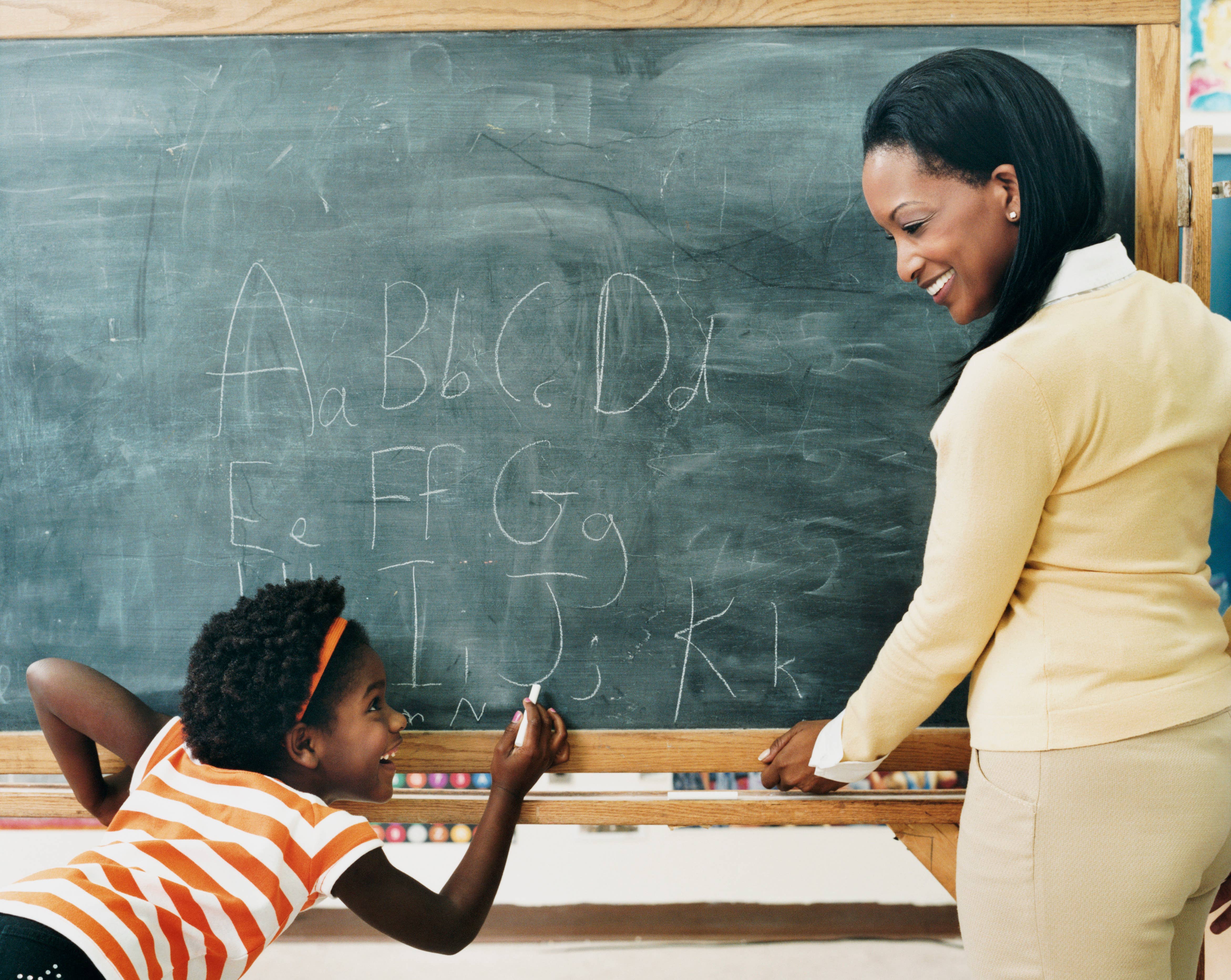 Black teacher. Parents sit and watch their children write on the blackboard.