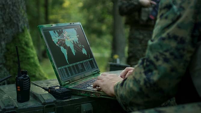 A Marine deep in the woods uses a laptop displaying world logistic data.