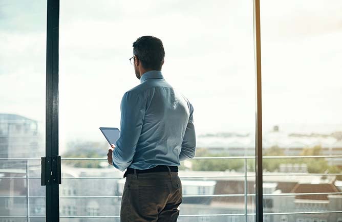 A businessman stands at a window overlooking the city while holding a tablet.