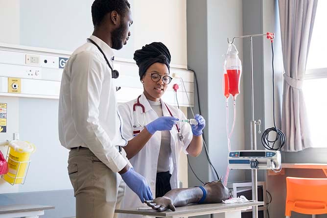 A medical doctor helps his colleague practice the method of injecting into an arm. They are working together in the hospital ward.