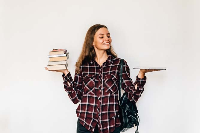 A girl balances books in one hand and a laptop in another.
