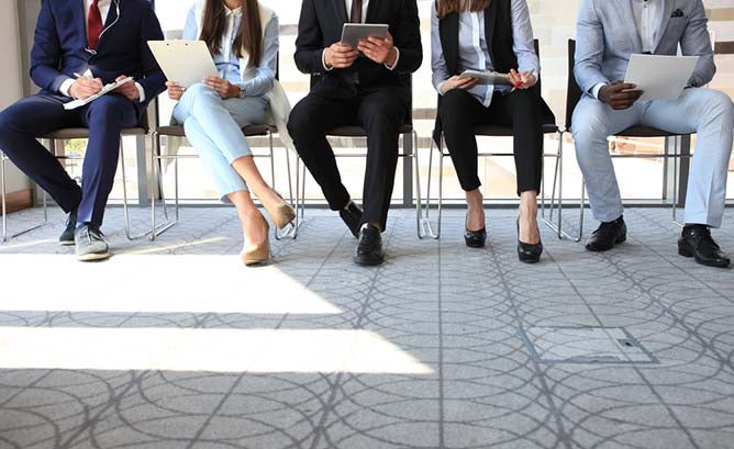 Five business people sit shoulder to shoulder, each reading a table or clipboard in their hands.