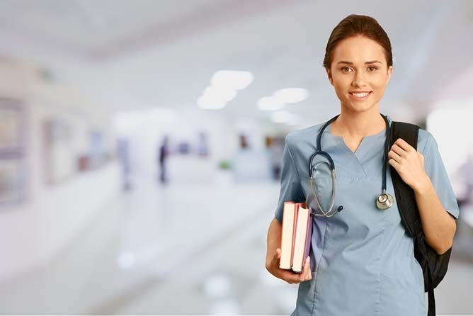A young nurse stands among a blurred background, holding two textbooks in her hand.