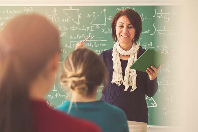A teacher lectures in front of her students with various mathematical equations scribble on the blackboard.