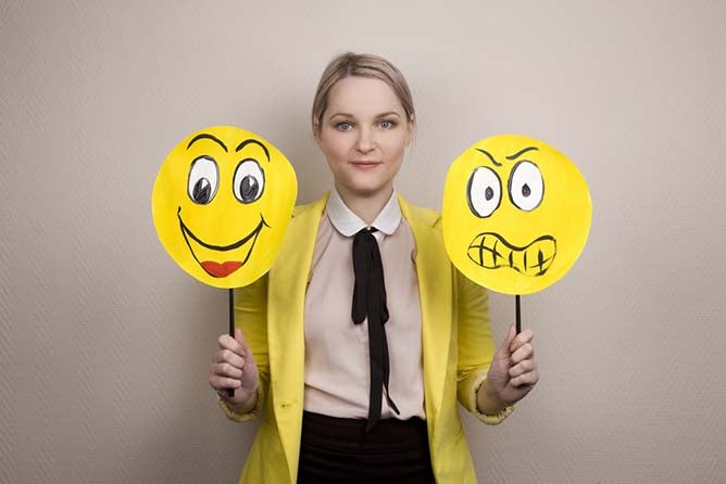 A young woman holds two cartoon faces; one happy, one sad.