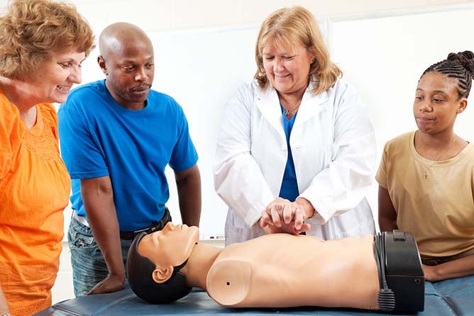 A group of students watch a doctor or nurse demonstrating CPR chest compressions on a dummy.