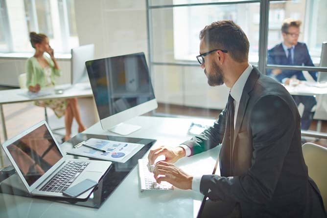 A businessman works at a desk with multiple devices in front of him.