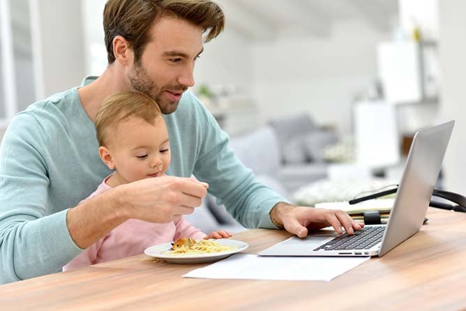 A man works on his laptop with his baby eating food on his lap.
