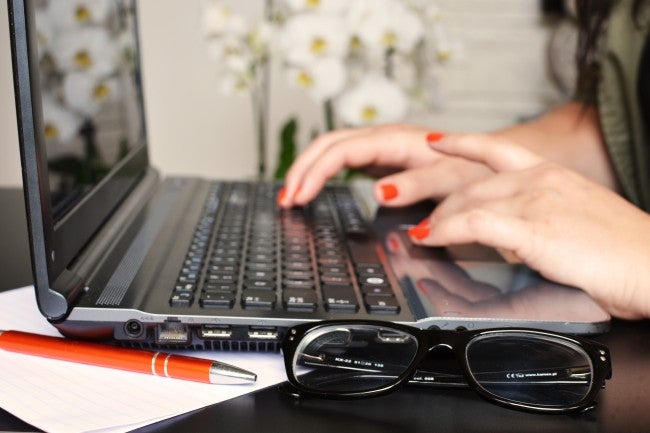 Woman typing on computer with pen and glasses nearby