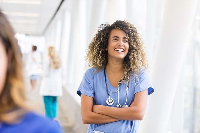 A smiling nurse stands with folded arms in a hallway.
