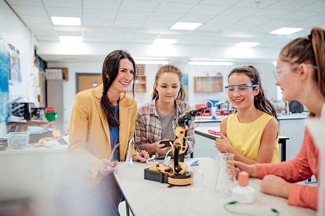 A group of girls operate a robotic arm under the supervision of their teacher.
