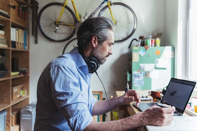 A middle-aged man works on a laptop in a cluttered backroom.