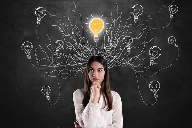 A woman stands in front of a blackboard with various lightbulbs drawn in chalk above her head.