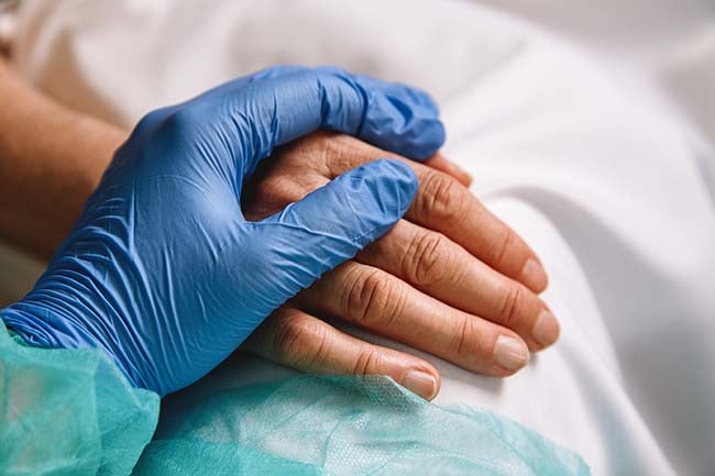 A closeup of a hand covered in a latex glove holding the hand of a patient laying in bed.