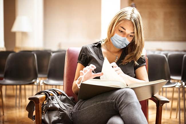 A young student studies a text book while wearing a mask.