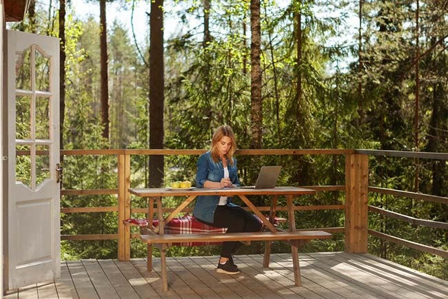 Young woman reading document and working on laptop computer sitting at table on wooden balcony of country house surrounded by pine trees