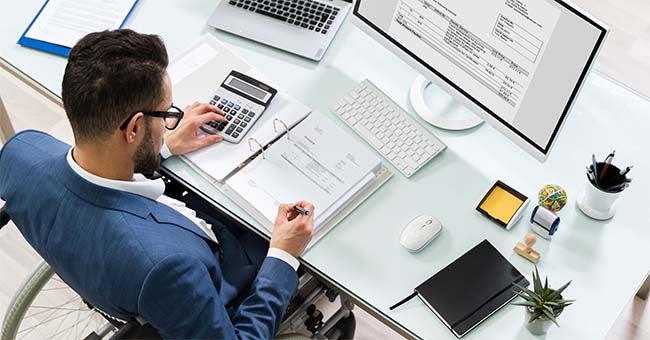 A businessman sitting at a computer types on a ten key and takes notes.