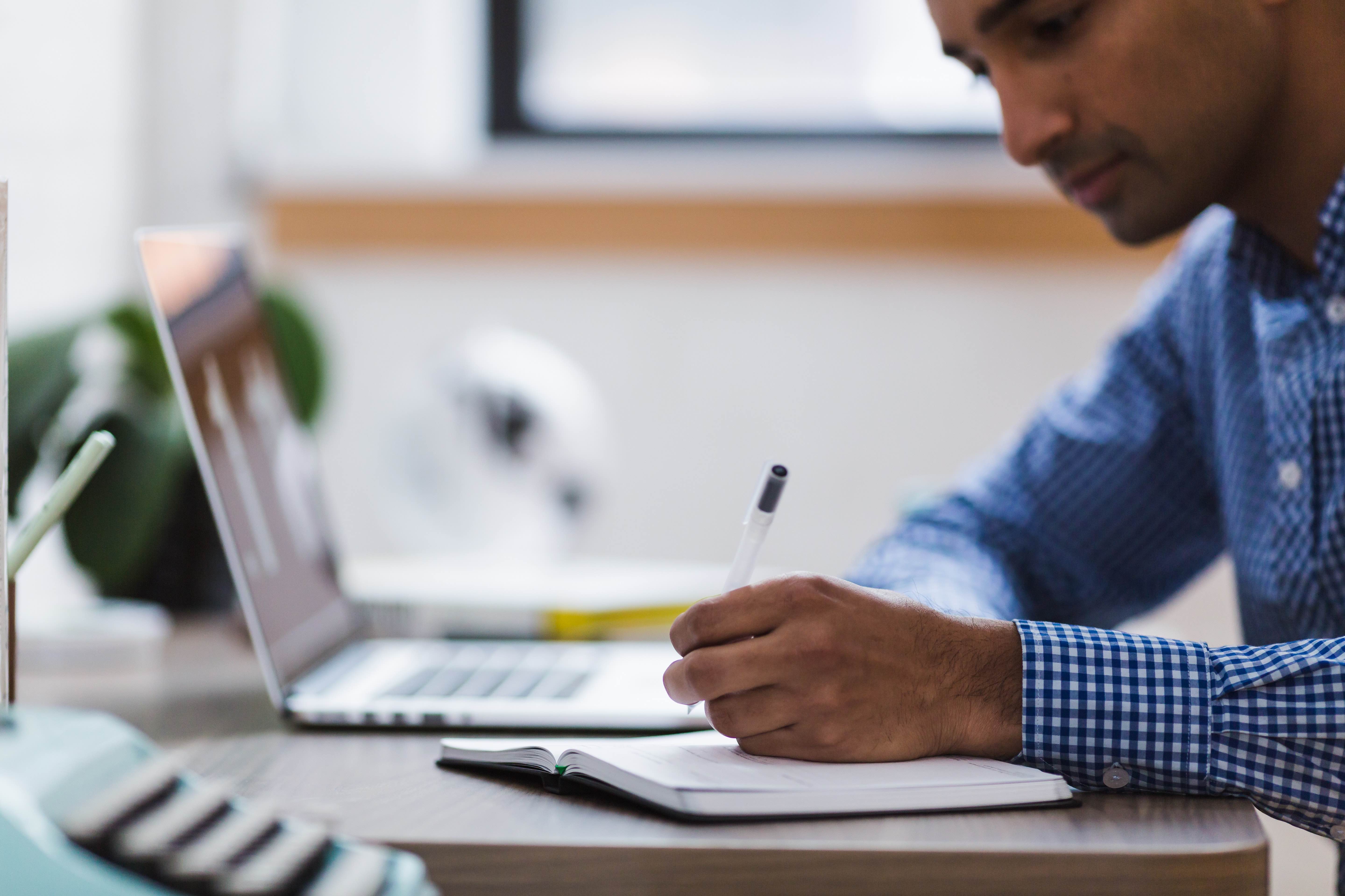 close up of a person writing in a notebook with a laptop open in front of them