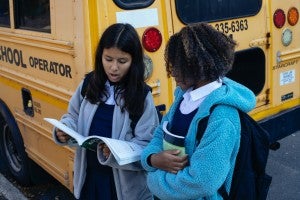 two girls reading a book by a school bus